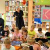 Dzieci siedzą na podłodze i przeglądają książki, za nimi stoi bibliotekarka i przygląda się grupie