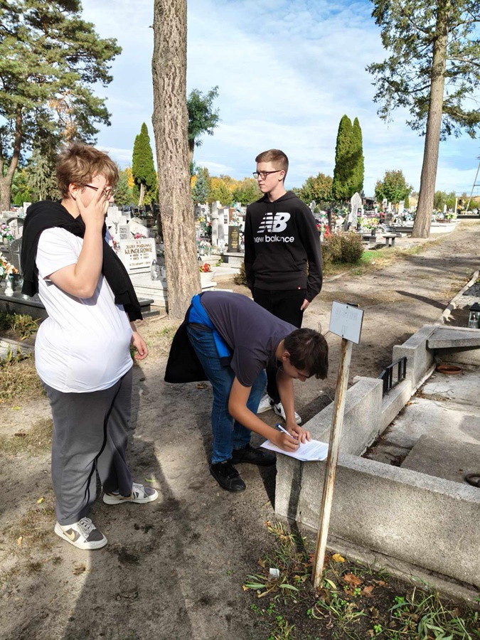 3 osoby stojące przed grobem na cmentarzu, jedna z nich zapisuje coś na kartce papieru