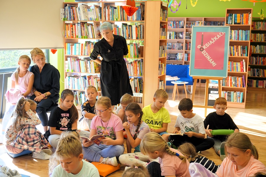 Dzieci siedzą na podłodze i przeglądają książki, za nimi stoi bibliotekarka i przygląda się grupie