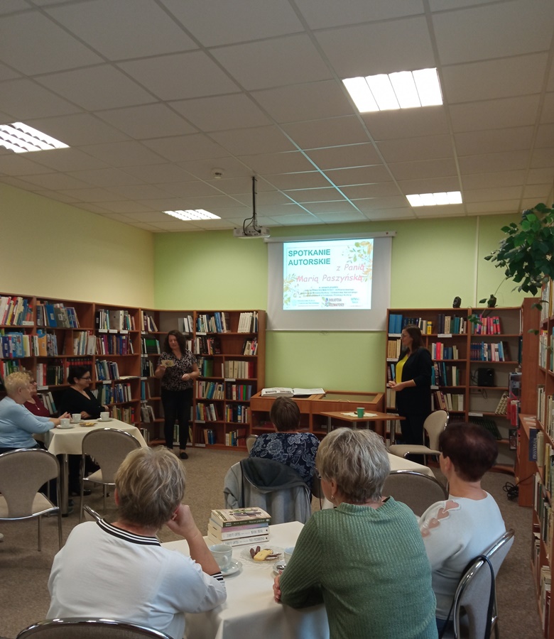 Widownia zebrana w czytelni, publiczność siedzi przy stolikach, przed nimi stoi zaproszona autorka i bibliotekarka która zapowiada gościa i rozpoczyna spotkanie