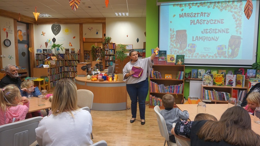 bibliotekarka stoi przed zebranymi na zajęciach uczestnikami, pokazuje im przykładowy wykonany lampion i tłumaczy co uczestnicy wykonają w czasie zajęć