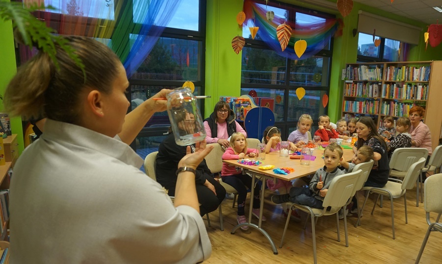 bibliotekarka stoi przed uczestnikami zajęć, trzyma w rękach szklany słoik i tłumaczy uczestnikom co bedą robić na zajęciach