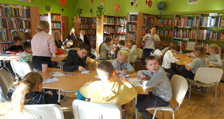 Dzieci uczestniczące w zajęciach siedzą przy stołach, bibliotekarka prowadząca lekcję mówi do zebranych uczestników