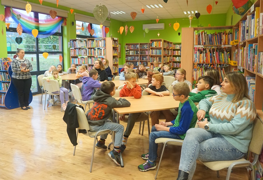 uczestnicy spotkania siedzą przy stołach, bibliotekarka prowadząca zajęcia stoi z boku i obserwuje uczestników