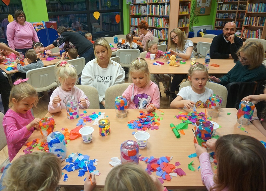 grupa uczestników razem z opiekunami siedzą przy stole i ozdabiają słoiki kolorowymi kawałkami papieru