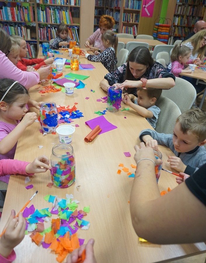 grupa uczestników razem z opiekunami siedzą przy stole i ozdabiają słoiki kolorowymi kawałkami papieru