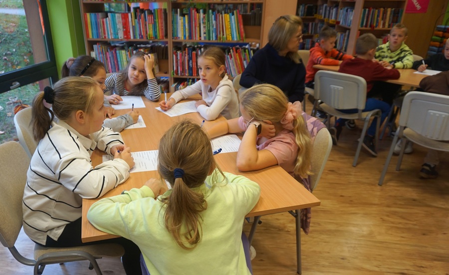 dzieci z opiekunami siedzą przy stolikach i piszą coś na kartkach papieru