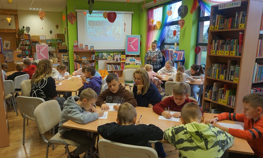 dzieci z opiekunami siedzą przy stolikach i piszą coś na kartkach papieru